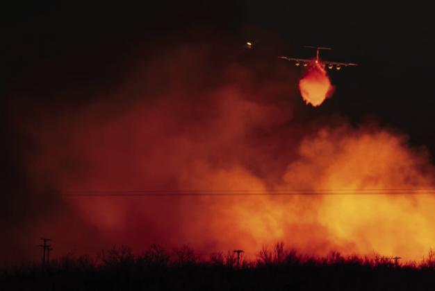 A Texas A&amp;M Forest Service plane drops water on a wall of flames that reaches into the sky as the Smokehouse Creek fire reignited o March 3 near Miami. | TEXAS A&amp;M AGRILIFE SAM CRAFT.